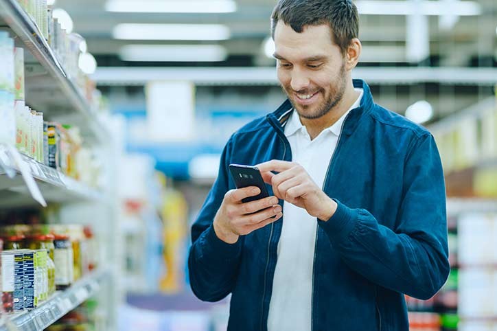 At the Supermarket: Handsome Man Uses Smartphone, Smiles while Standing at the Canned Goods Section. 
