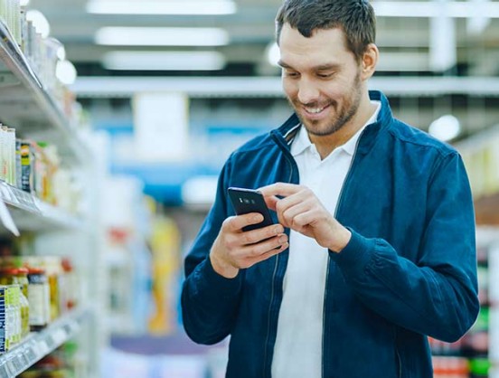 At the Supermarket: Handsome Man Uses Smartphone, Smiles while Standing at the Canned Goods Section. 
